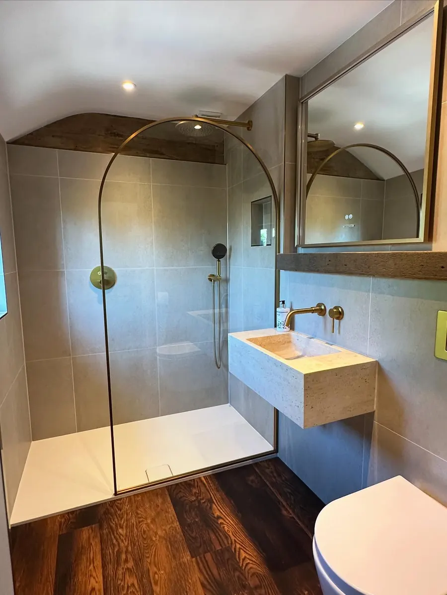 A shower room with grey tiles, a stone trough basin, brass tap and arched shower screen