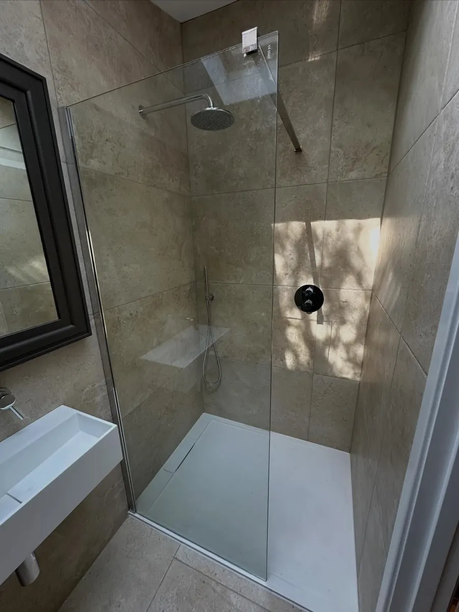A walk-in shower with a glass screen, black rain shower head and travertine-style tiles