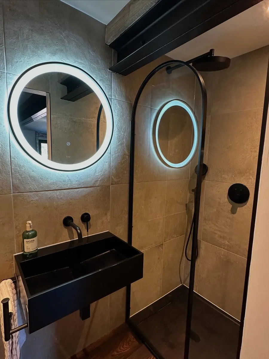 A bathroom with a black basin tap, black shower and round LED mirror